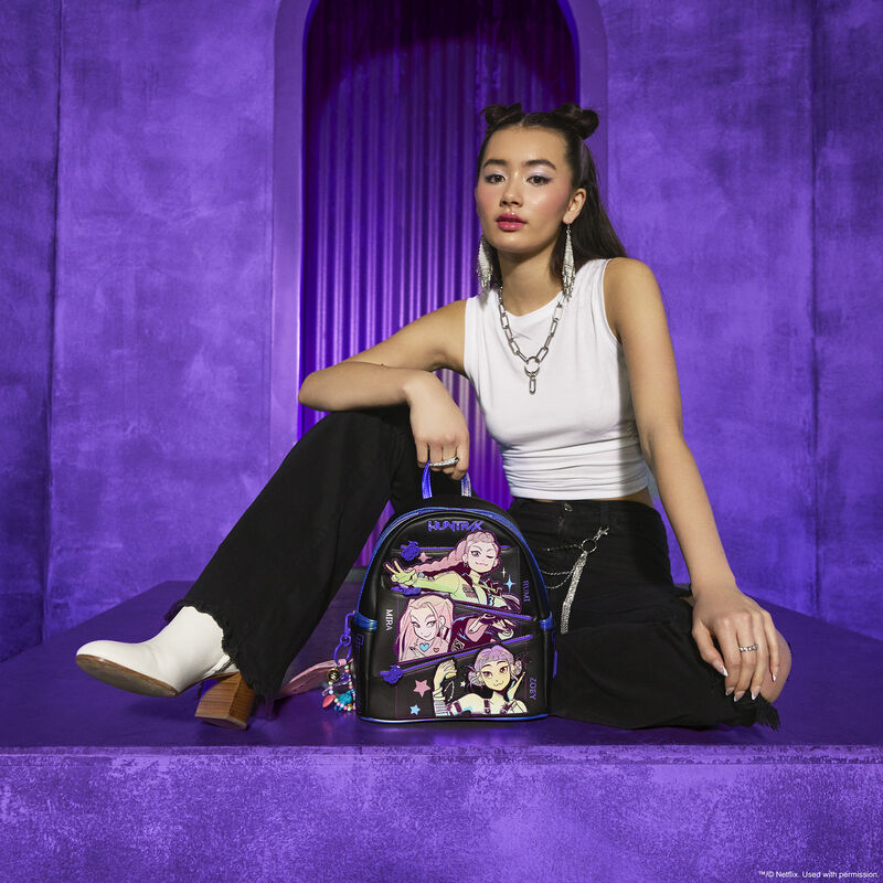 Woman wearing a white top, black pants, and white boots, sitting behind the Loungefly HUNTR/X mini backpack. Everything around her is purple. 